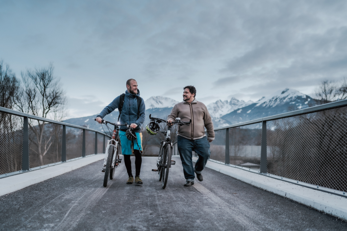 zwei Männer warm angesogen laufen ihr Fahrad schiebend über Fahrradbrücke und reden