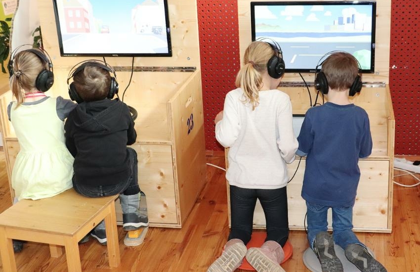 4 Kinder sichtbar von hinten, sie knien und sitzen vor Holzstationen der Klimaversum-Wanderausstellung, und schauen auf Bildschirme.