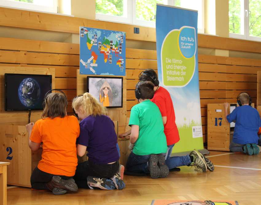 Kinder sitzen vor zwei Stationen mit Bildschirmen der Klimaversum-Wanderausstellung auf ihren Knien. Man sieht sie von hinten.