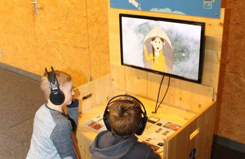 Zwei Kinder mit Kopfhörern mit Kopfhörern sitzen vor einem Bildschirm, auf dem eine Zeichentrick-Figur zu ihnen spricht.