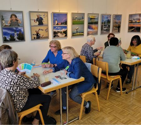 Je vier Senior:innen sitzen an zwei Tischen, Karten in der Handj, hinter ihnen Bilder an der Wand.