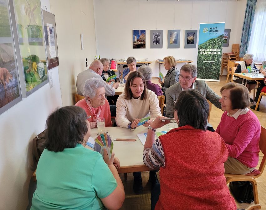 Senior:innen sitzen mit einer Mitarbeiterin vom Klimabündnis Steiermark an Tischen, sie deutet auf Karten auf ihrer Hand.