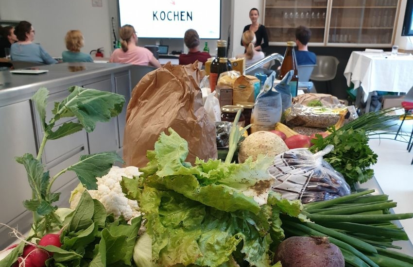 Salat, Frühlingszwiebel, Käferbohnen und weiteres Gemüse liegen auf einer Küchenzeile. Im Hintergrund eine Person im Stehen auf die sitzenden Personen schauen