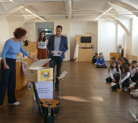 Cornelia Schweiner öffnet den Deckel einer Holzkiste die auf einem Sitzwürfel steht. Darhinter zwei Personen. Rechts davon sitzen Kinder auf dem Boden und schauen zu.