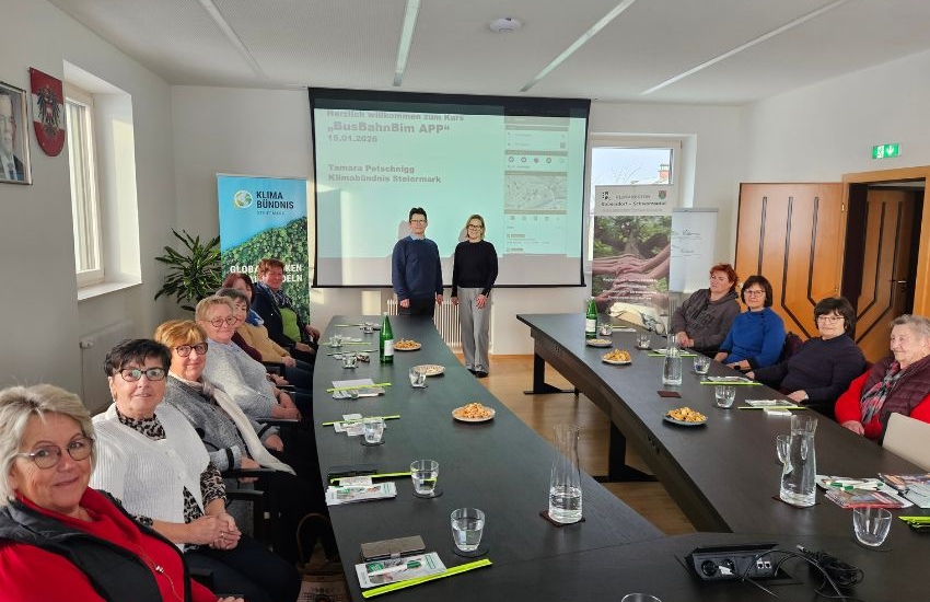Senioren sitzen an langer Tafel und lächeln, im Hintergrund Beamer und Vortragende