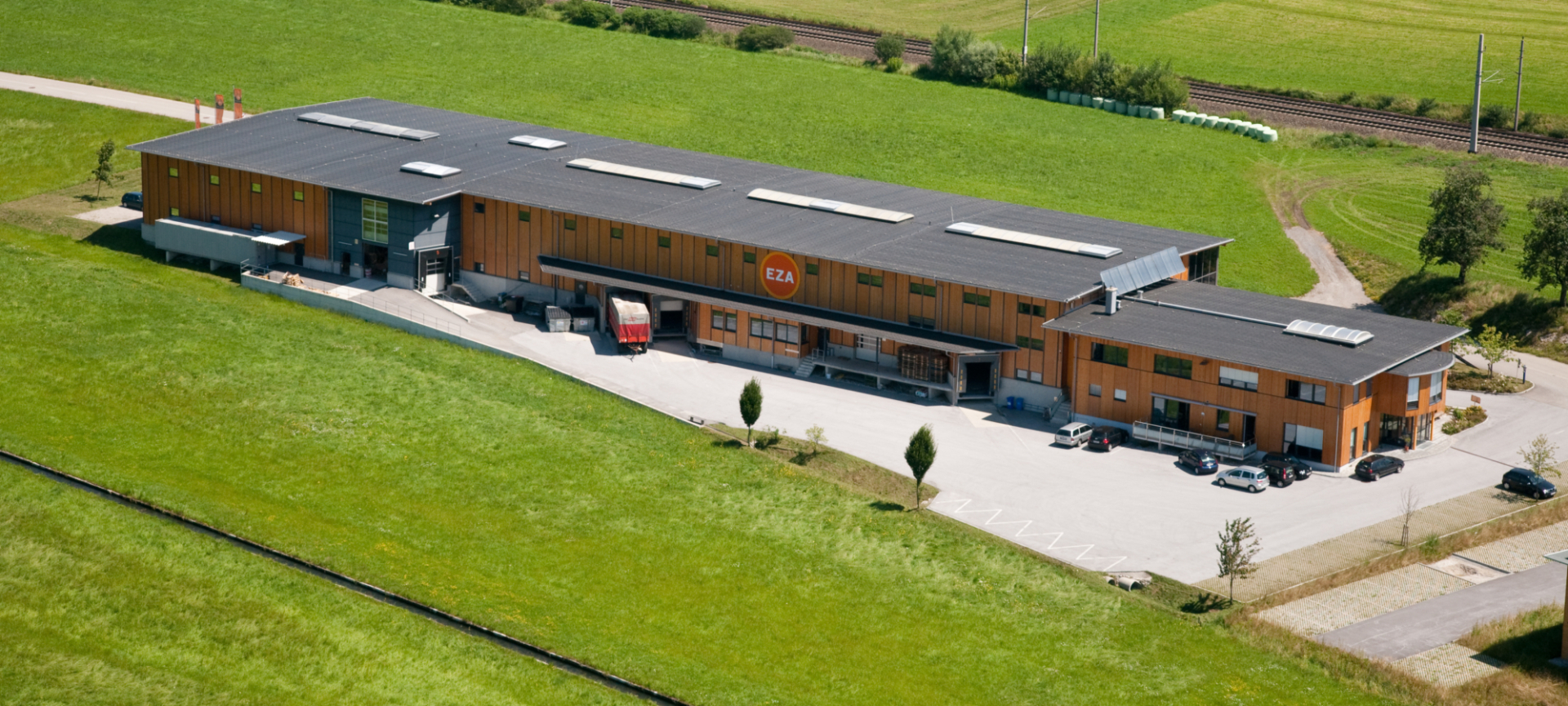 Luftaufnahme eines langgestreckten Betriebsgebäudes in ländlicher Umgebung, umgeben von grünen Feldern