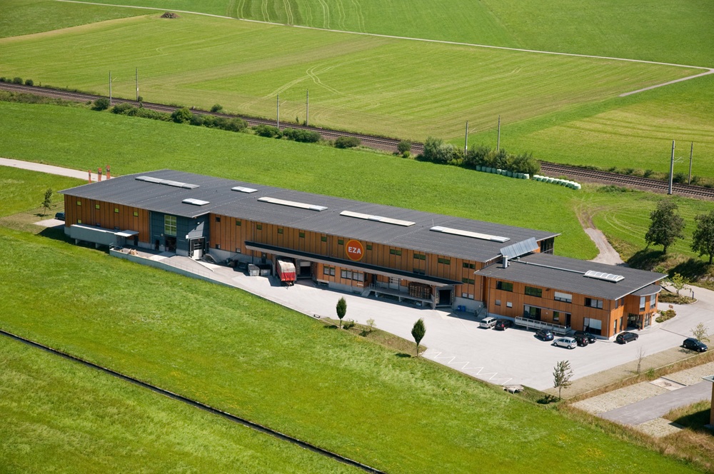 Luftaufnahme eines langgestreckten Betriebsgebäudes in ländlicher Umgebung, umgeben von grünen Feldern