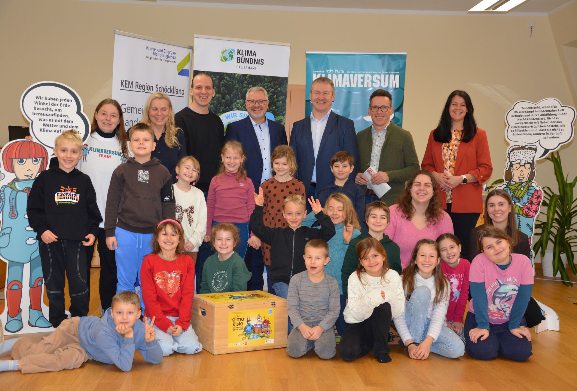 Gruppenfoto von 15 Kindern im Volksschulalter und 7 Erwachsenen mit der Klimakiste und den Pappfiguren der Ausstellung