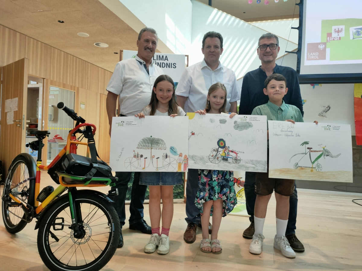 Drei Kinder zeigen ihre Zeichnungen von Fahrrädern, im Hintergrund drei Männer.